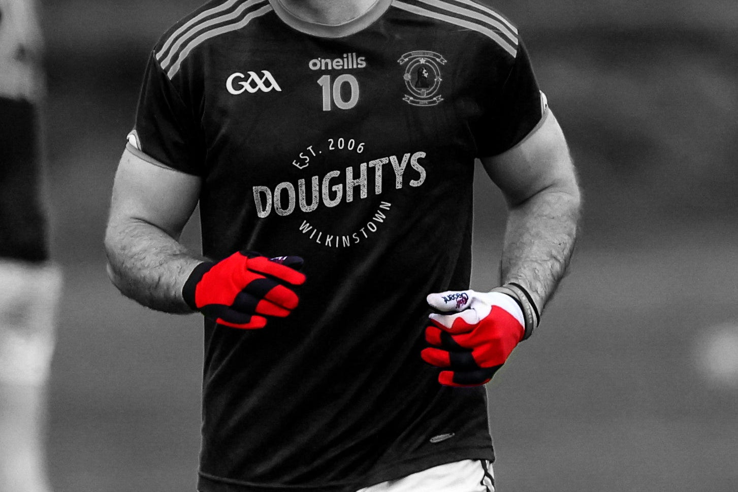 A man wearing a black Cork football jersey with white shorts and red gloves, captured in a black and white photo with the red gloves and jersey color highlighted. Visible brands include O'Neill, CCA, and Doughtys.